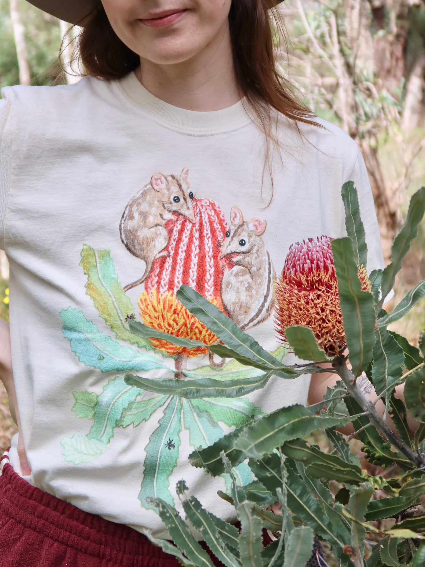 Person wearing a white t-shirt with a design of two honey possums on a banksia, standing in a natural setting.