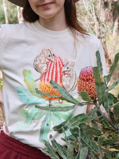 Person wearing a white t-shirt with a design of two honey possums on a banksia, standing in a natural setting.
